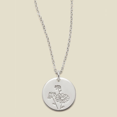 A silver chain necklace with a round silver disc pendant featuring an engraved daisy flower design, displayed against a solid light beige background.