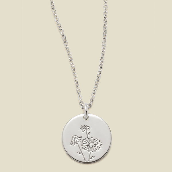 A silver chain necklace with a round silver disc pendant featuring an engraved daisy flower design, displayed against a solid light beige background.