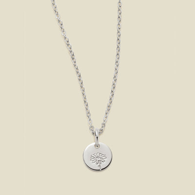 A silver chain necklace with a round silver disc pendant featuring an engraved daisy flower design, displayed against a solid light beige background.