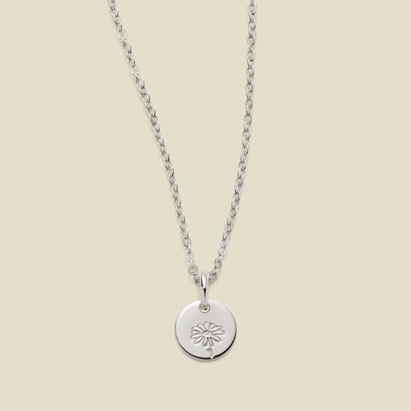 A silver chain necklace with a round silver disc pendant featuring an engraved daisy flower design, displayed against a solid light beige background.
