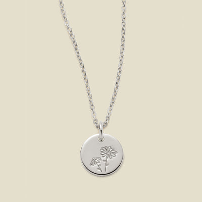 A silver chain necklace with a round silver disc pendant featuring an engraved daisy flower design, displayed against a solid light beige background.