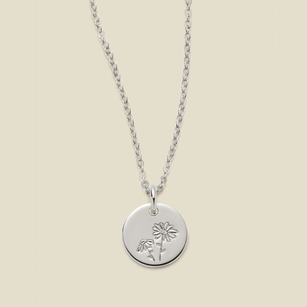A silver chain necklace with a round silver disc pendant featuring an engraved daisy flower design, displayed against a solid light beige background.
