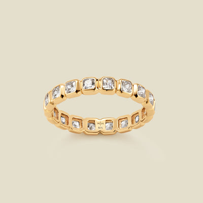 A gold eternity ring featuring a continuous row of square cut clear gemstones, shown on a solid light beige background.