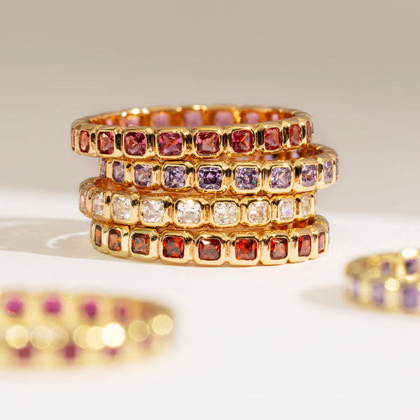 A stack of four gold eternity rings, each with either dark red, purple, clear, or orange-red square cut gemstones in a single continuous row, shown on a warm white background with other rings blurred in the foreground.