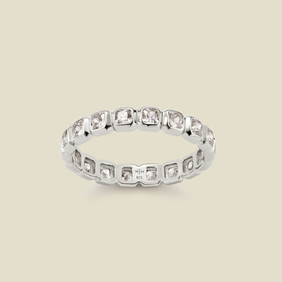 A silver eternity ring featuring a continuous row of square cut clear gemstones, shown on a solid light beige background.