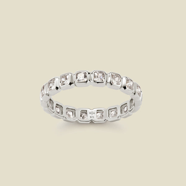 A silver eternity ring featuring a continuous row of square cut clear gemstones, shown on a solid light beige background.