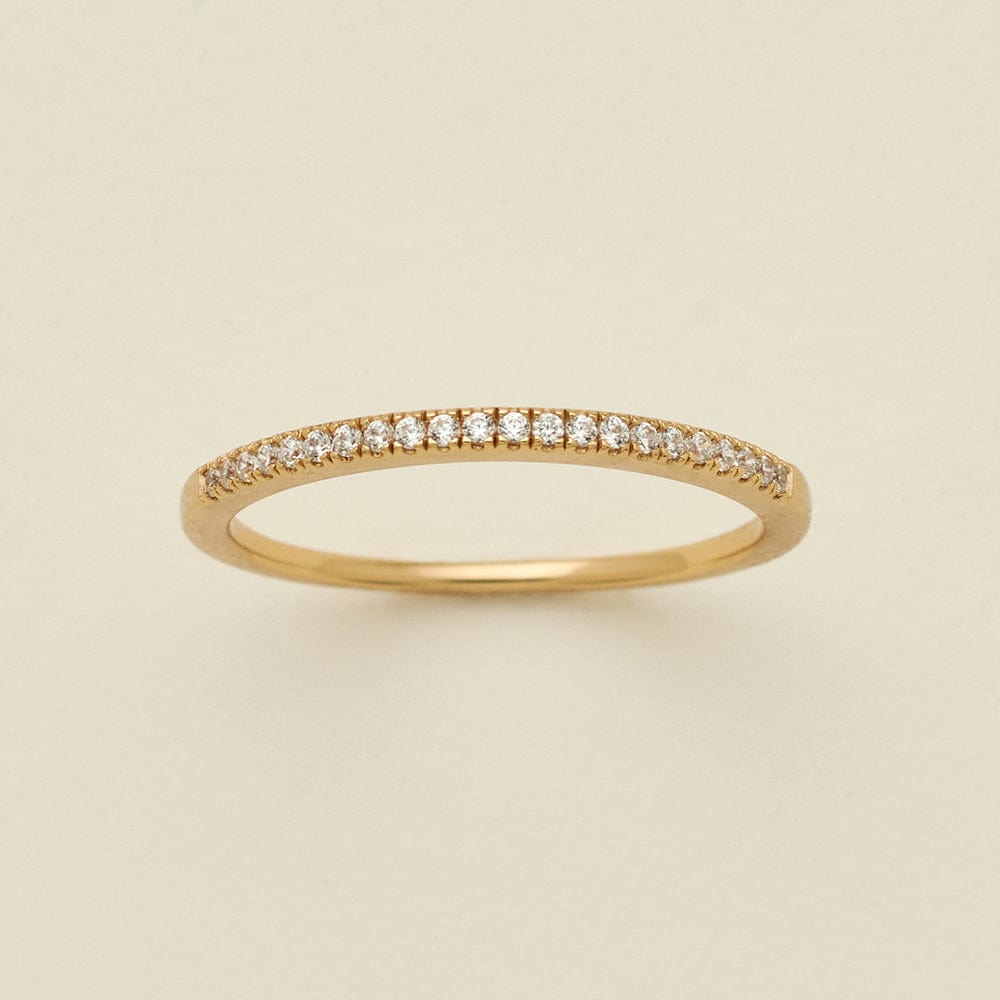 A gold ring featuring a single row of small round-cut clear gemstones, photographed on a clean, solid, warm off-white background.