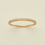 A gold ring featuring a single row of small round-cut clear gemstones, photographed on a clean, solid, warm off-white background.