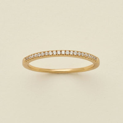 A gold ring featuring a single row of small round-cut clear gemstones, photographed on a clean, solid, warm off-white background.