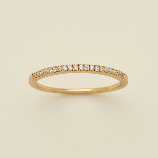 A gold ring featuring a single row of small round-cut clear gemstones, photographed on a clean, solid, warm off-white background.