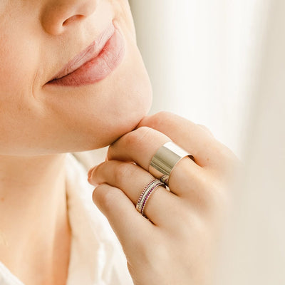 A close-up of a person's hand resting against their chin, showcasing a thick polished gold cigar band ring and a stack of three thin gold rings, each with either pink, clear, or red gemstones placed in a single row across the front.