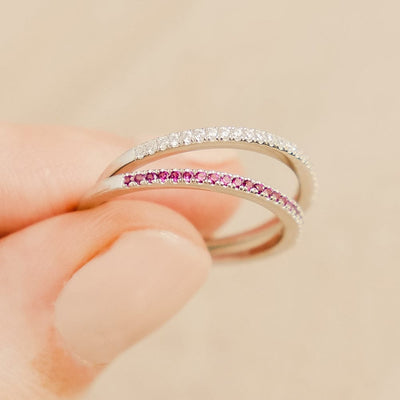 A close-up of two fingers holding thin silver rings, each with a single row of small gemstones across the front. One has clear gemstones, and the other has red.
