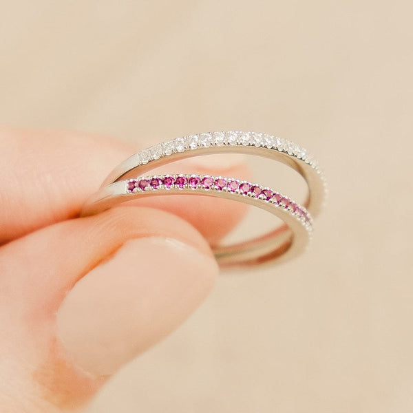 A close-up of two fingers holding thin silver rings, each with a single row of small gemstones across the front. One has clear gemstones, and the other has red.