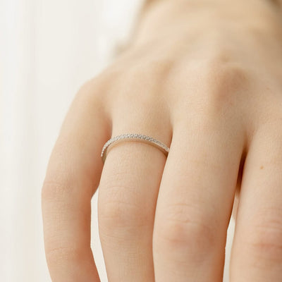 A close-up of a hand wearing a thin silver ring, featuring a single row of small round-cut clear gemstones, set against a bright white blurred background.