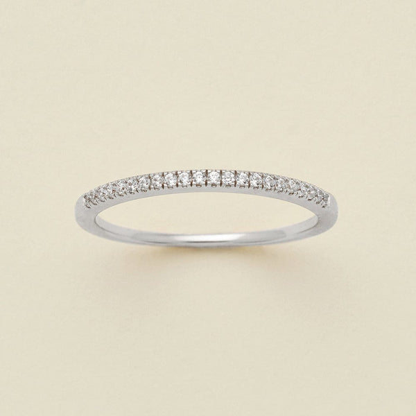 A silver ring featuring a single row of small round-cut clear gemstones, photographed on a clean, solid, warm off-white background.