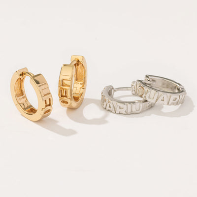 A pair of gold huggie hoops with the word 'LEO' in raised letters on the front, sitting upright next to a silver pair of the same hoops with the word 'AQUARIUS' in raised lettering. 