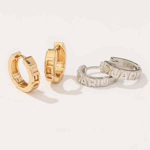 A pair of gold huggie hoops with the word 'LEO' in raised letters on the front, sitting upright next to a silver pair of the same hoops with the word 'AQUARIUS' in raised lettering. 