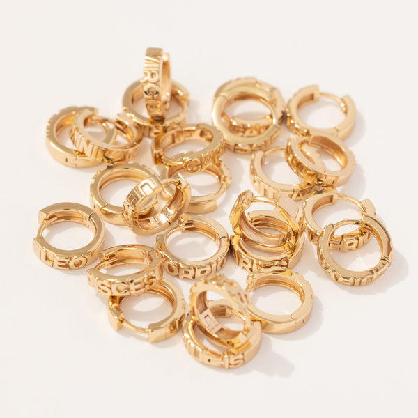 Multiple gold huggie hoops scattered on a solid bright white surface, each with the name of a zodiac sign across the front in raised letters.
