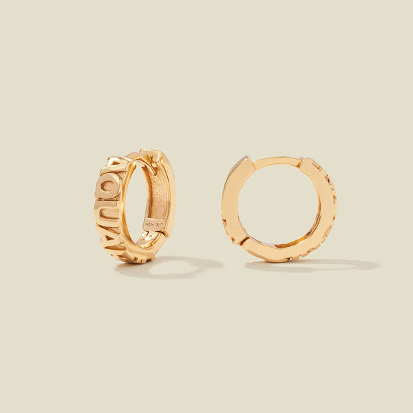 A pair of gold huggie hoop earrings on a light beige background. One earring is shown from the side, displaying a polished outer surface, while the other is shown from the front, showcasing the word 'AQUARIUS' in raised letters.