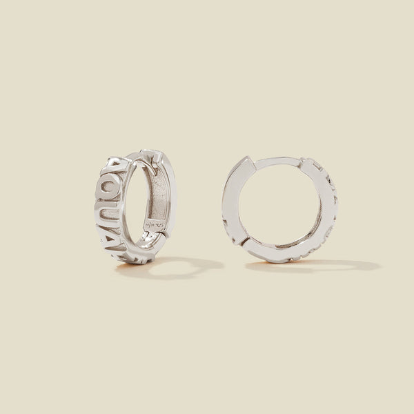 A pair of silver huggie hoop earrings on a light beige background. One earring is shown from the side, displaying a polished outer surface, while the other is shown from the front, showcasing the word 'AQUARIUS' in raised letters.