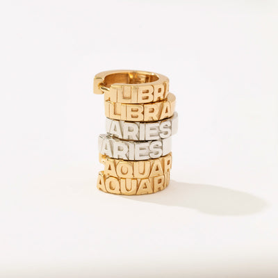 A stack of three pairs of huggie hoop earrings, two gold and one silver. Each of the earrings has the name of a zodiac sign in raised letters along the front.