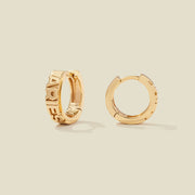 A pair of gold huggie hoop earrings on a light beige background. One earring is shown from the side, displaying a polished outer surface, while the other is shown from the front, showcasing the word 'ARIES' in raised letters.
