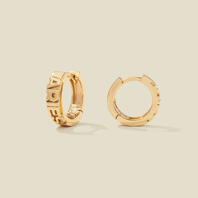 A pair of gold huggie hoop earrings on a light beige background. One earring is shown from the side, displaying a polished outer surface, while the other is shown from the front, showcasing the word 'ARIES' in raised letters.