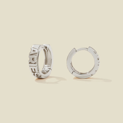 A pair of silver huggie hoop earrings on a light beige background. One earring is shown from the side, displaying a polished outer surface, while the other is shown from the front, showcasing the word 'ARIES' in raised letters.