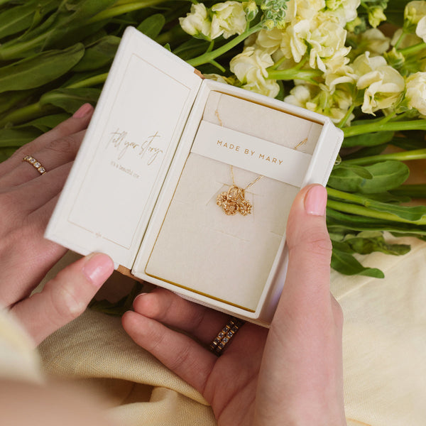 A cream colored jewelry box sitting open with a gold pendant chain necklace holding three flower charms, shown in front of a bouquet of white flowers.