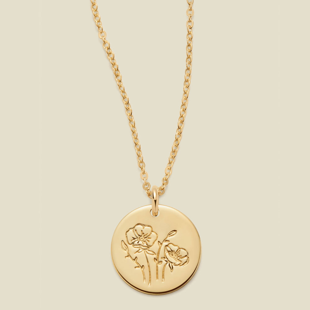 A gold chain necklace with a round gold disc pendant featuring an engraved poppy flower design, displayed against a solid light beige background.