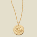 A gold chain necklace with a round gold disc pendant featuring an engraved poppy flower design, displayed against a solid light beige background.
