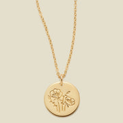 A gold chain necklace with a round gold disc pendant featuring an engraved poppy flower design, displayed against a solid light beige background.