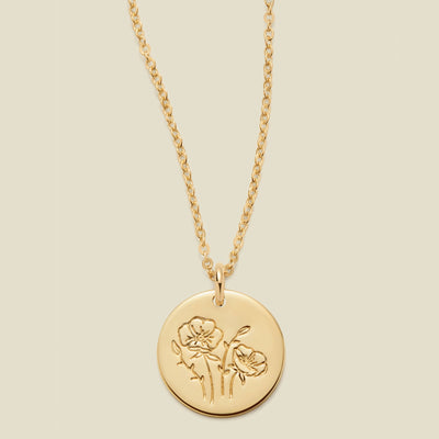 A gold chain necklace with a round gold disc pendant featuring an engraved poppy flower design, displayed against a solid light beige background.