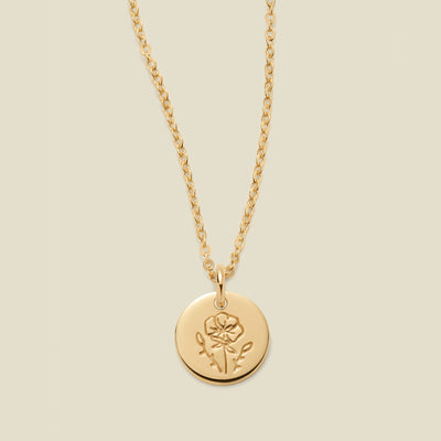 A gold chain necklace with a round gold disc pendant featuring an engraved poppy flower design, displayed against a solid light beige background.