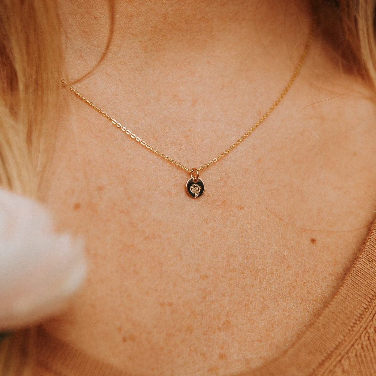 A close-up of a person in a brown ribbed top wearing a delicate gold pendant chain with a flat round disc charm. The disc is engraved with a poppy flower design.