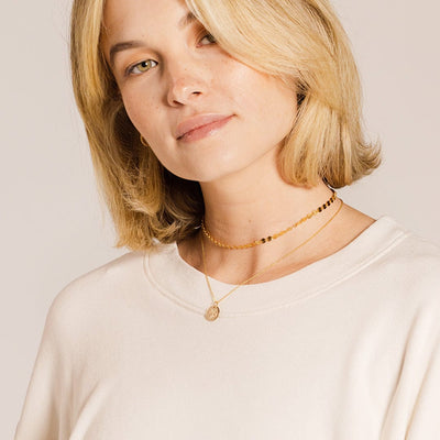 A blonde woman in a white long sleeve top wearing a gold choker made up of flat round discs linked together, and a longer delicate pendant chain with a flat round disc charm. The charm is engraved with a floral design.