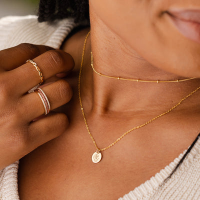 A close-up of a person in a cream knit sweater wearing a dainty gold choker with stationary round beads and a delicate pendant chain with a flat round disc charm. The charm is engraved with a poppy flower design.
