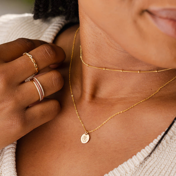 A close-up of a person in a cream knit sweater wearing a dainty gold choker with stationary round beads and a delicate pendant chain with a flat round disc charm. The charm is engraved with a poppy flower design.