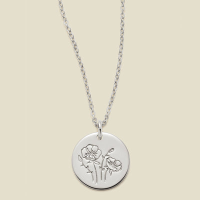 A silver chain necklace with a round silver disc pendant featuring an engraved poppy flower design, displayed against a solid light beige background.