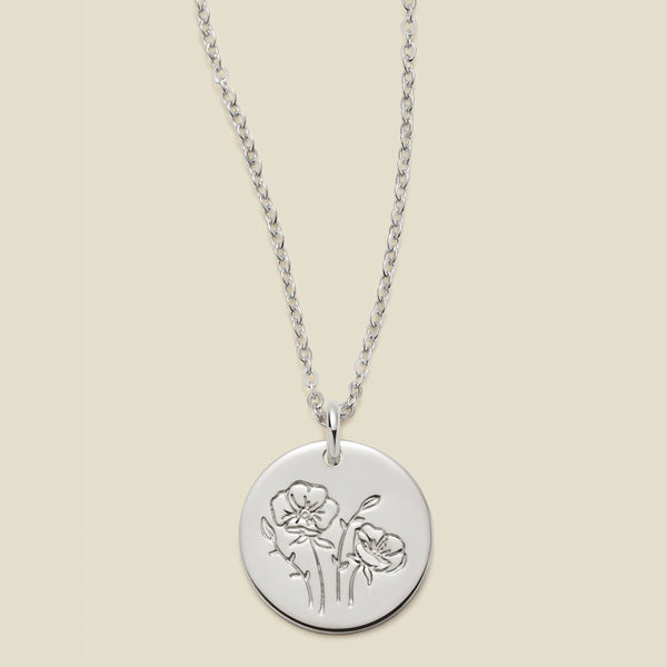 A silver chain necklace with a round silver disc pendant featuring an engraved poppy flower design, displayed against a solid light beige background.