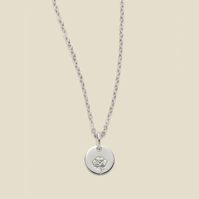 A silver chain necklace with a round silver disc pendant featuring an engraved poppy flower design, displayed against a solid light beige background.
