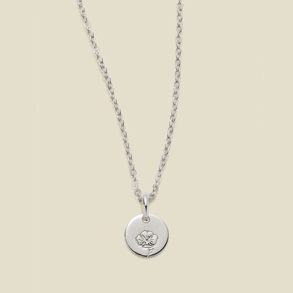 A silver chain necklace with a round silver disc pendant featuring an engraved poppy flower design, displayed against a solid light beige background.