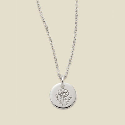 A silver chain necklace with a round silver disc pendant featuring an engraved poppy flower design, displayed against a solid light beige background.