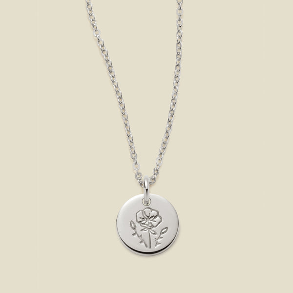 A silver chain necklace with a round silver disc pendant featuring an engraved poppy flower design, displayed against a solid light beige background.