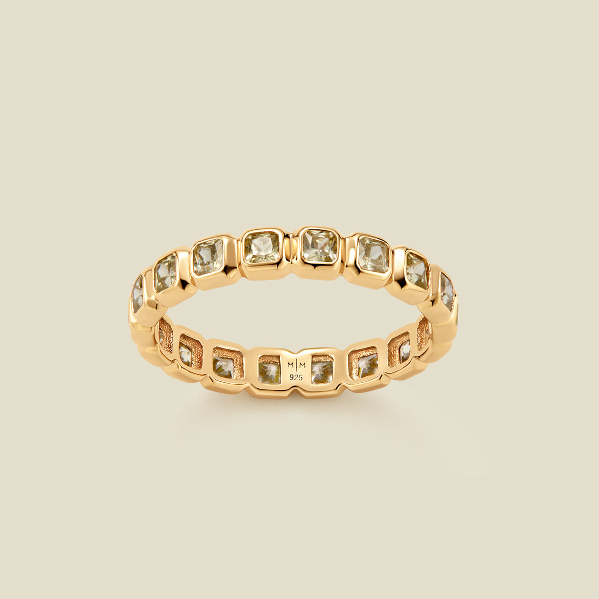 A gold eternity ring featuring a continuous row of square cut light green gemstones, shown on a solid light beige background.