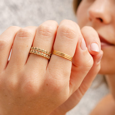 A close-up of a hand wearing three stacked thin gold band rings and two stacked gold eternity style rings, each with a single continuous row of square cut light green gemstones.
