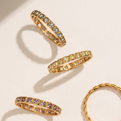 Four gold eternity rings, three on their sides showcasing a single continuous row of square cut gemstones in the colors light blue, light green, and purple, set against a white background with natural shadows.