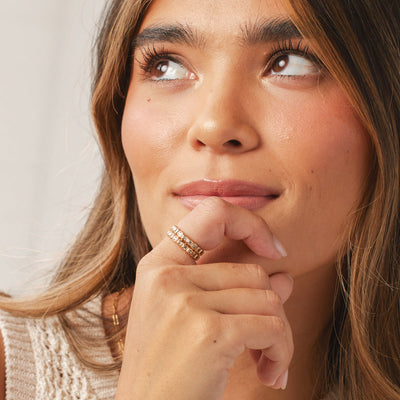 A person holding their hand up to their chin showing two stacked gold eternity rings, each with a continuous row of light green square cut gemstones, set against a white background.