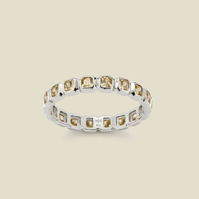 A silver eternity ring featuring a continuous row of square cut light green gemstones, shown on a solid light beige background.