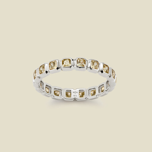 A silver eternity ring featuring a continuous row of square cut light green gemstones, shown on a solid light beige background.